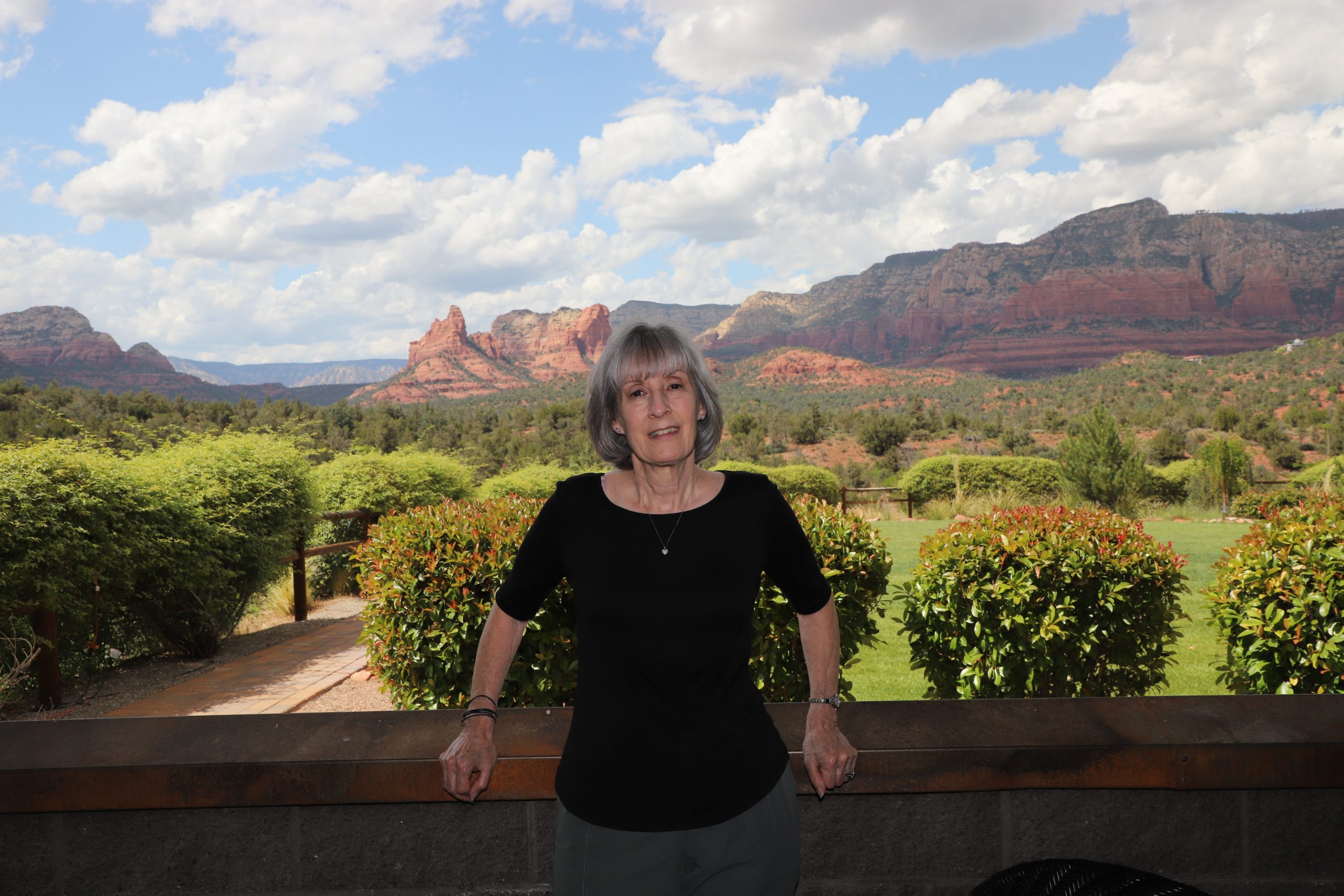 A woman with short gray hair, wearing a black shirt, stands leaning against a stone wall. Behind her is a landscape with green bushes, red rock formations, and a partly cloudy sky.